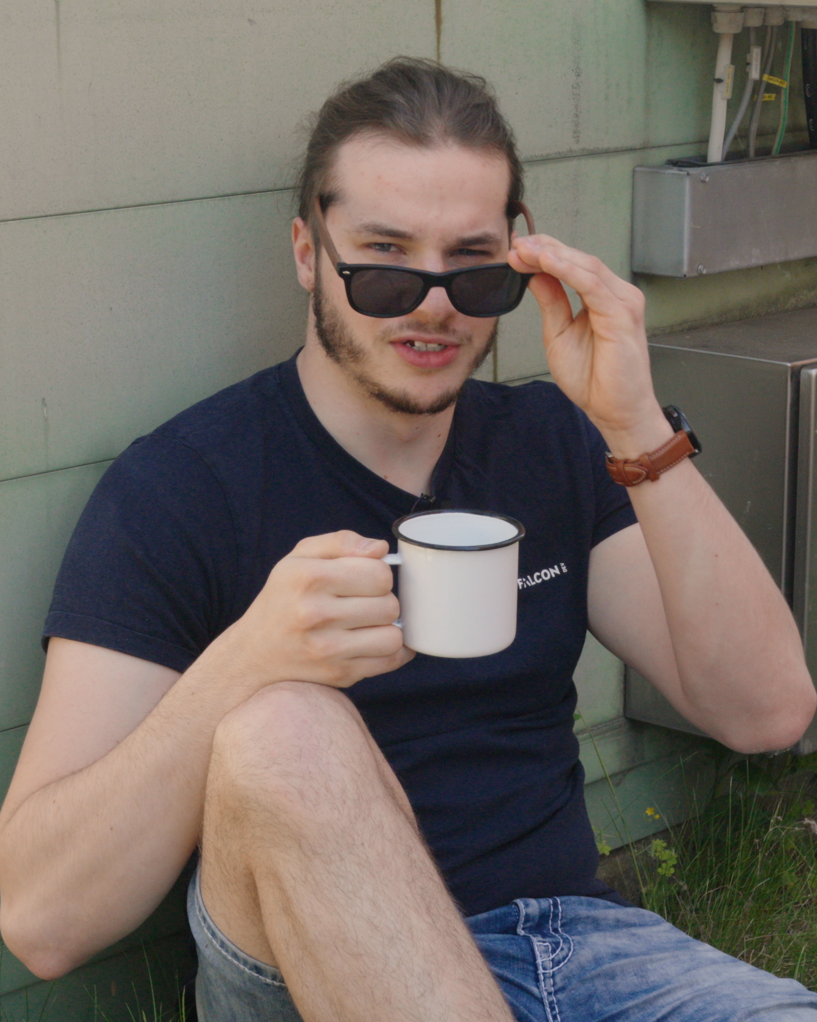 Julian sitzt angelehnt an eine Hauswand mit einem Kaffee in der Hand und schiebt seine Sonnenbrille mit einem fragenden Gesichtsausdruck herunter