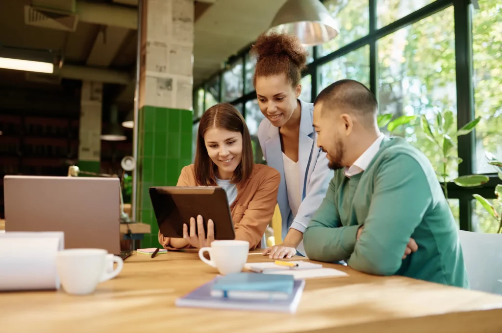 Drei Personen arbeiten gemeinsam an einem Holztisch in einem hellen, modernen Büroraum mit großen Fenstern. Eine Person hält ein Tablet in der Hand, die anderen beiden schauen darauf. Laptops, Notebooks und Kaffeetassen stehen auf dem Tisch, während sie ihre Eigenentwicklung für das Kundenportal besprechen.