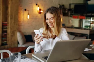 Junge Geschäftsfrau liest eine Nachricht auf einem Smartphone mit einem Laptop vor sich