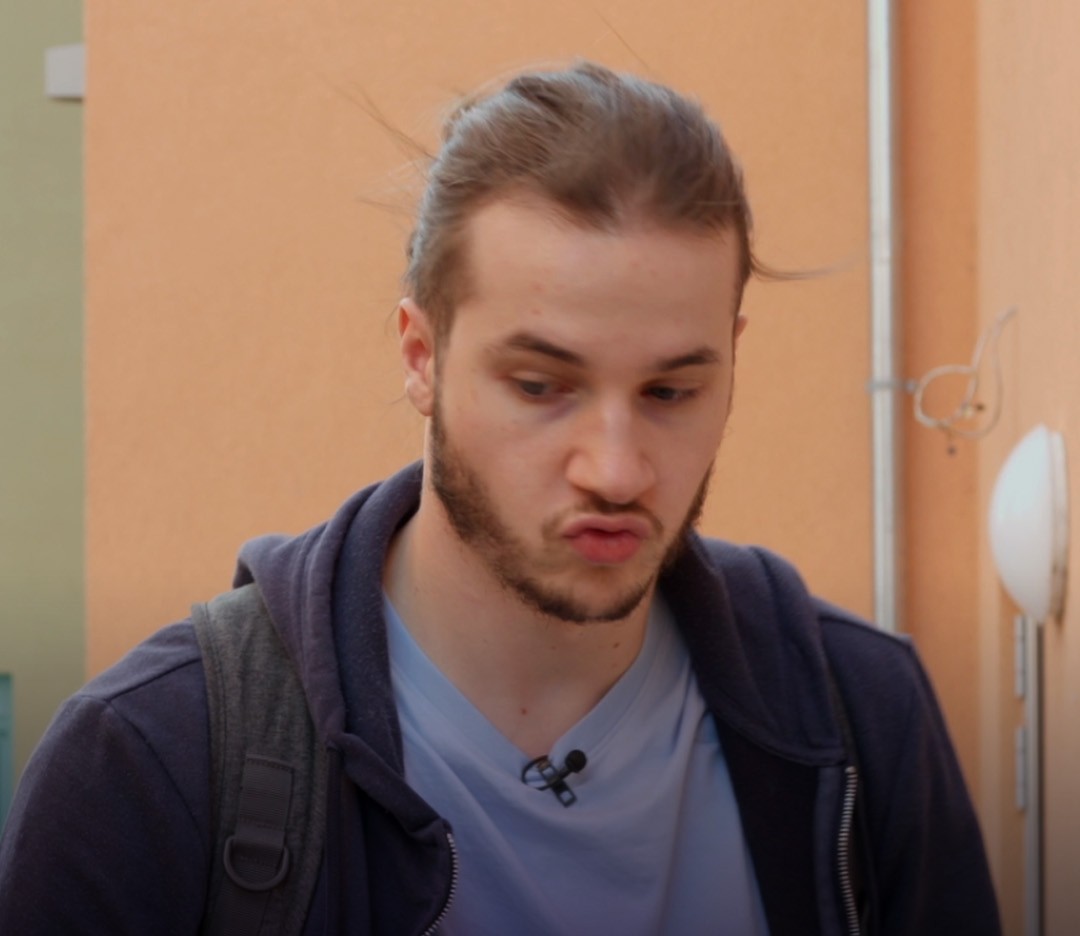 Junger Mann mit kurzem, glattem Haar und Bart, der in einem orangefarbenen Innenhof steht. Er trägt ein blaues T-Shirt und eine graue Jacke. Im Hintergrund sind Parkett und eine Lampe sichtbar. Der Mann wirkt nachdenklich oder konzentriert.