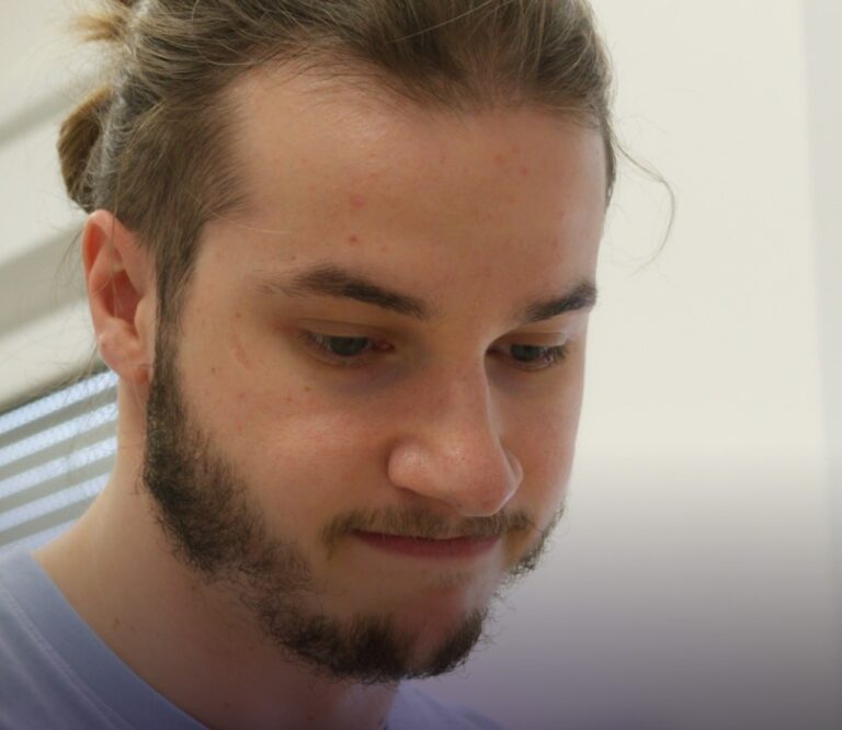 Junger Mann mit dünnem Bart und langen Haaren, der konzentriert auf ein Dokument schaut. Hintergrund mit neutraler Farbgebung.